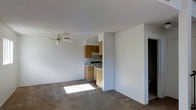 This two-floor residential space offers a straightforward and functional layout designed for comfortable everyday living. The main living area is open and airy, featuring neutral-toned carpet flooring that extends throughout, creating a cohesive and inviting atmosphere. A ceiling fan is centrally positioned overhead, providing both lighting and air circulation. Natural light filters into the living space, brightening the interior and enhancing the sense of openness. The kitchen is situated adjacent to the living area in an efficient galley-style arrangement, featuring warm wood-toned cabinetry, an electric range with overhead ventilation, and a under-counter refrigerator. The white countertops and appliances contribute to a clean, practical aesthetic. The textured ceiling adds a classic residential character to the space. A doorway visible from the main living area leads to additional rooms, suggesting a well-connected floor plan across both levels. The overall ambiance is clean, neutral, and move-in ready, offering a blank canvas that lends itself well to personalization. The two-floor layout provides added separation between living and private spaces, making this a well-rounded and versatile residential unit.