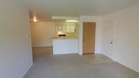 This single-floor unit is designed with an open-concept layout that seamlessly connects the living area and kitchen, creating a cohesive and practical living space. The living area is finished with wall-to-wall carpeting in a neutral beige tone, lending a soft and understated feel to the space.

The kitchen is positioned centrally and defined by a built-in peninsula that doubles as a breakfast bar, providing both additional prep space and a natural divide between the cooking and living zones. White cabinetry lines the kitchen walls, offering ample storage, while under-cabinet lighting and a flush-mounted fluorescent fixture illuminate the workspace effectively. A stove is visible along the back wall, and the kitchen flooring transitions to a hard surface, distinguishing it from the carpeted living area.

The entry area is located adjacent to the kitchen, featuring a solid wood-tone door that provides a clean and functional entry point. The walls throughout are finished in a clean white, contributing to a bright and open atmosphere. Overhead lighting is present in both the kitchen and living areas. The overall design is simple, practical, and move-in ready.