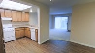 This single-floor residence offers a functional and open layout designed for comfortable everyday living. The kitchen is equipped with warm honey-toned wood cabinetry, a granite-look countertop, a built-in sink, and a white dishwasher. A skylight above the kitchen brings in an added touch of natural light, brightening the workspace. The kitchen flows seamlessly into the adjacent living area through an open pass-through design, making the space feel connected and airy. Durable wood-style hard flooring runs through the kitchen and transitional areas, providing a clean and modern aesthetic. The living area features plush carpeting underfoot, offering a comfortable contrast to the hard flooring. A large sliding glass door fitted with vertical blinds anchors the living space, allowing abundant natural light to filter in while providing access to the exterior. The neutral wall tones throughout create a calm, versatile backdrop suitable for a variety of interior styles. The overall layout is straightforward and practical, with a smooth transition between the cooking and living zones, making this a well-organized and inviting single-level home.