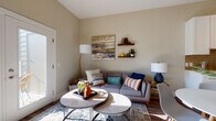 This single-floor residence is thoughtfully designed with a cohesive, contemporary aesthetic throughout. The open-concept kitchen serves as a functional centerpiece, featuring white shaker cabinetry extending to the upper walls, stainless steel appliances including a gas range, microwave, and refrigerator, a tile backsplash, and an extended island with quartz countertops ideal for dining or food preparation. Warm wood-look flooring runs through the kitchen and connects seamlessly to the living and dining areas.

The living area is bright and welcoming, with warm neutral wall tones, floating wood shelves for display, and a door providing direct access to a private balcony, filling the space with natural light. The bedroom is generously proportioned, showcasing large windows with views to the outdoors, soft carpeting, and elevated ceiling height that adds an airy quality to the room. A striking four-poster bed frame anchors the space.

The bathroom is finished with white shaker-style cabinetry, a wide vanity with a clean white countertop, a large mirror, and wood-look flooring that ties the space to the rest of the unit. Overall, the residence presents a polished, modern living environment with consistent finishes and abundant natural light throughout.