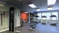 This fitness center offers a comprehensive workout environment designed to accommodate a range of exercise needs. The space is finished with durable rubber flooring, providing a safe and practical surface throughout. Overhead, flush-mounted panel lighting ensures the area is consistently and evenly illuminated, complemented by natural light streaming in through windows along one wall.

The layout is thoughtfully organized into distinct zones. The cardio area features multiple treadmills positioned near the windows, allowing users to exercise with a view of the surrounding greenery. A central open area provides space for free-weight and bench work, equipped with an adjustable workout bench.

Toward the back of the space, a bold orange accent wall defines a secondary training zone, housing a weight bench and additional strength equipment. Along the near wall, a multi-function cable machine offers a wide variety of resistance-based exercises. The cool gray tones of the walls throughout the main area create a focused, energizing atmosphere. A structural column is incorporated neatly into the layout, dividing the zones without disrupting the overall flow. The space presents a well-rounded fitness facility that is both practical and inviting.