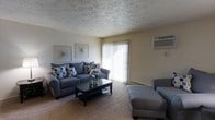 This single-floor space centers around a living room designed for comfort and functionality. The room features wall-to-wall carpeting in a neutral tone, creating a warm foundation that complements the light-colored walls. A textured ceiling adds subtle visual interest overhead.

Natural light enters through a large sliding glass door dressed with vertical blinds, offering access to the outdoors and brightening the interior. A wall-mounted air conditioning unit is positioned nearby, ensuring year-round climate control.

The living area is generously sized, accommodating a sectional-style sofa in slate blue, accented with patterned throw pillows, alongside a chaise lounge chair in a matching tone. A dark-framed rectangular coffee table anchors the seating arrangement, providing practical surface space. Two framed abstract artworks adorn the wall above the sofa, adding a decorative touch. The overall ambiance is casual and inviting, with a neutral color palette throughout that lends itself well to a variety of furnishing styles and personal tastes.