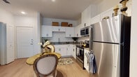 This single-floor residence offers a thoughtfully designed layout that combines functionality with modern style. The open kitchen is a standout feature, showcasing crisp white shaker-style cabinetry with matte black hardware, dark countertops, and a full suite of stainless steel appliances including a refrigerator, electric range, microwave, and dishwasher. Light wood-style flooring runs throughout, lending warmth and continuity to the space. A compact dining area with a round table sits adjacent to the kitchen, creating a seamless flow between cooking and dining. The living area, visible in the floor plan, provides a comfortable gathering space with room for a full sofa arrangement. The bedroom is serene and well-sized, featuring neutral tones, and direct access to a spacious walk-in closet with built-in shelving and hanging rods. The bathroom is finished with a wide vanity topped with a light stone-look countertop, an undermount sink, and a tub-shower combination with a decorative curtain. Recessed lighting and ample ceiling height contribute to a bright, airy ambiance throughout the residence. The overall design balances contemporary finishes with a welcoming, comfortable character.