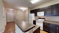 This single-floor residence at Parks At Vine offers a thoughtfully designed layout that balances open communal spaces with comfortable private areas. The kitchen serves as a central highlight, featuring rich dark espresso cabinetry with brushed nickel hardware, neutral-toned countertops, wood-look flooring, and white appliances including a refrigerator, electric range, and microwave. A peninsula with additional cabinetry and counter space provides convenient workspace and a natural transition into the living area. The open living room is carpeted and includes a ceiling fan with light fixture, offering a bright, airy feel enhanced by French doors that invite natural light. The bedrooms continue the carpeted aesthetic and are equipped with ceiling fans, multiple windows with blinds for natural light and privacy, and walk-in closet space outfitted with wire shelving systems. The bathrooms are finished with dark vanity cabinetry, tile flooring, a large mirror, and a tub-shower combination, maintaining the consistent neutral color palette found throughout. Overall, the residence presents a clean, functional ambiance with a cohesive design scheme.