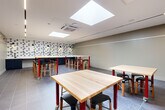 Located at 728 Broadway in Saugus, Massachusetts, this single-floor makers space is thoughtfully designed to support collaborative, hands-on work. The open-plan layout provides a generous, unobstructed working environment filled with natural light from multiple large skylights recessed into the white ceiling. Supplementing the natural light, sleek linear LED fixtures run along the ceiling perimeter, ensuring consistent, well-distributed illumination throughout the space.

The flooring consists of large-format dark grey tiles that lend a clean, industrial character to the room. Multiple butcher-block work tables with bold red legs on casters are arranged throughout, offering flexibility for reconfiguration. Black padded stools accompany each workstation, providing comfortable seating for extended use.

A standout design feature is the geometric black-and-white triangle-patterned accent wall at one end of the space, which adds visual energy and creative character. This wall also incorporates adjustable black task lighting and a built-in dark cabinetry station with a sink, supporting a variety of project types. A wall-mounted cassette-style HVAC unit ensures climate control year-round. Neutral grey walls on the remaining sides keep the atmosphere clean and focused. The space is accessed via a standard door with an illuminated exit sign, meeting all safety requirements.