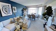 This single-floor space showcases a thoughtfully designed open-concept layout that seamlessly connects the living and dining areas into one cohesive, inviting environment. The most striking design element is the deep teal-blue accent wall, which serves as a bold backdrop to the living area and adds a sense of depth and sophistication to the space.

The living area features plush, light-colored carpeting that extends throughout, contributing to a warm and comfortable atmosphere. A neutral-toned sofa and ottoman are complemented by decorative touches, including a striking floral arrangement in the foreground that adds a fresh, organic element to the interior.

Built-in open shelving along the blue accent wall provides functional display space for decorative items and greenery, adding personality and visual interest. The dining area is furnished with a round wooden table surrounded by elegant striped upholstered chairs, creating an intimate setting for gatherings.

Large bay-style windows dressed with patterned sheer curtains allow generous amounts of natural light to flood the space, enhancing its open and airy feel. The overall ambiance is polished and curated, balancing bold design choices with comfortable, everyday livability.