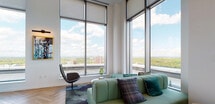 Located at 5 Livingston Ave in New Brunswick, NJ, this single-floor lounge space combines bold design with elevated functionality. Floor-to-ceiling curtain-wall windows dominate the seating area, flooding the interior with natural light and offering sweeping panoramic views of the surrounding cityscape and landscape. The herringbone-patterned hardwood flooring adds a refined, classic texture that contrasts beautifully with the contemporary furnishings throughout.

The lounge seating area features plush, teal-upholstered sofas accented with decorative throw pillows, arranged atop richly toned area rugs that anchor the space. A sculptural swivel chair provides additional seating while complementing the modern aesthetic. The open layout allows for flexible social arrangements and encourages natural movement through the space.

The bar area is the visual centerpiece of the room, defined by a dark stone-tiled counter, patterned bar stools, and vivid teal lacquered walls. Backlit open shelving displays an curated collection of geode specimens and decorative objects, creating a gallery-like effect. Above, a sculptural brass multi-globe chandelier casts warm ambient light, enhancing the dramatic, jewel-toned atmosphere. Together, these elements create a cohesive, upscale environment suited for social gatherings.