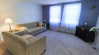 This single-floor space centers around a well-appointed living room that combines classic architectural elements with a welcoming atmosphere. The room is finished with wall-to-wall carpeting in a neutral tone, providing a soft and comfortable foundation throughout the space.

A striking blue-purple accent wall anchors the room, creating a bold contrast against the lighter surrounding walls, adding depth and visual interest to the interior. Two large windows dressed with sheer white curtains allow an abundance of natural light to filter into the space, lending the room an airy, bright quality. The windows also highlight a classic cast-iron radiator beneath them, a functional and characterful feature that speaks to the building's traditional construction.

The living area is generously proportioned, comfortably accommodating an L-shaped sectional sofa arrangement in a warm taupe upholstery, accented with decorative pillows. A small side table with a decorative floral accent completes the seating area. A television setup is positioned to one side of the room, making the space practical for everyday living.

Overall, the space offers a balance of classic details and comfortable, functional living in a bright, inviting setting.