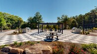 Located at 1665 Wesleyan Dr in Macon, Georgia, this outdoor amenity space is a well-appointed gathering area designed for relaxation and community engagement. At its centerpiece sits a striking black-and-white checkered tile patio, which anchors the space with a bold, graphic aesthetic. A sleek modern pergola with a dark metal frame rises above the patio, providing shade and structure for seating arrangements beneath it. Adjacent to the patio, a built-in grill station offers an ideal setup for outdoor cooking and entertaining. Winding gravel pathways connect the various zones of the space, guiding visitors through carefully curated planted beds filled with ornamental shrubs, flowering plants, young trees, and natural mulch borders. A dedicated fire pit area, surrounded by Adirondack-style chairs and accented with decorative gravel, provides a cozy retreat for cooler evenings. String lights are strung between tall posts throughout the grounds, lending a warm, inviting ambiance as the day transitions to evening. The perimeter is enclosed by a chain-link fence, beyond which mature Georgia pines and hardwoods create a natural green backdrop. Lush lawn areas and well-maintained landscaping throughout reinforce the space's polished, community-oriented character under open blue skies.