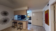 This single-floor residence offers a practical and functional layout across its main living areas. The kitchen is designed in a galley style, equipped with dark-finish cabinetry, a gas cooktop, black appliances including a full-size refrigerator and microwave, and a double sink with ample counter space. A peninsula with a built-in breakfast bar extends from the kitchen, providing seating for two bar stools and serving as a natural divider between the kitchen and the adjacent living area. Overhead cabinetry with under-cabinet lighting adds both storage and illumination to the workspace. Tile flooring runs through the kitchen area, transitioning to carpet in the living and sleeping spaces. The living room features a ceiling fan with light fixture, a window fitted with horizontal blinds for light control, and enough space to accommodate a large sectional sofa. The bedroom is carpeted and includes a ceiling fan, a window with blinds, and sufficient space for a full bedroom suite. The bathroom features a tub and shower combination enclosed by sliding glass doors with chrome hardware. Textured ceilings are consistent throughout the unit, contributing to its classic residential character.