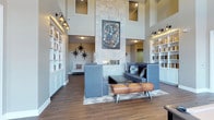This single-floor space makes an immediate impression with its dramatically high ceilings, which flood the interior with natural light through clerestory windows positioned near the roofline. The focal point of the room is a floor-to-ceiling stone fireplace wall, clad in light-toned tile and adorned with a framed piece of artwork, lending both visual weight and artistic character to the space.

Flanking the room on both sides are floor-to-ceiling built-in bookshelves finished in crisp white, providing generous display and storage space while reinforcing a sense of symmetry throughout. Wall-mounted industrial-style sconce lighting illuminates the shelving, adding a warm, layered glow to the interior.

The seating area is thoughtfully arranged with contemporary upholstered furniture in muted blue-gray tones, complemented by a distinctive leather pommel-horse bench that serves as a unique decorative accent. Star-shaped wall décor punctuates the neutral walls with bold graphic detail. The wood-look tile flooring grounds the space with warmth, while a geometric area rug defines the central seating zone, contributing to the overall modern yet eclectic ambiance.