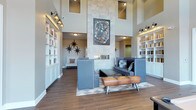 This single-floor space makes an immediate impression with its dramatically high ceilings, which flood the interior with natural light through clerestory windows positioned near the roofline. The focal point of the room is a floor-to-ceiling stone fireplace wall, clad in light-toned tile and adorned with a framed piece of artwork, lending both visual weight and artistic character to the space.

Flanking the room on both sides are floor-to-ceiling built-in bookshelves finished in crisp white, providing generous display and storage space while reinforcing a sense of symmetry throughout. Wall-mounted industrial-style sconce lighting illuminates the shelving, adding a warm, layered glow to the interior.

The seating area is thoughtfully arranged with contemporary upholstered furniture in muted blue-gray tones, complemented by a distinctive leather pommel-horse bench that serves as a unique decorative accent. Star-shaped wall décor punctuates the neutral walls with bold graphic detail. The wood-look tile flooring grounds the space with warmth, while a geometric area rug defines the central seating zone, contributing to the overall modern yet eclectic ambiance.