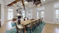 This elegantly designed single-floor space combines open-concept living with refined architectural details. Soaring vaulted ceilings with prominent dark wood beams define the interior, creating a dramatic sense of height and volume throughout. Light hardwood flooring flows seamlessly across the entire space, unifying its distinct functional zones.

The lounge area is anchored by a cluster of contemporary grey upholstered swivel chairs arranged around a tufted ottoman. A generous built-in window bench, fitted with teal accent cushions, lines one wall beneath a row of large windows fitted with roller shades, allowing for adjustable natural light. French doors with transom windows provide direct outdoor access and further brighten the room.

The dining area features a substantial reclaimed wood table surrounded by upholstered dining chairs, set atop a vibrant teal patterned area rug. Statement woven pendant lights hang overhead, complementing the vaulted ceiling. A fireplace and decorative wall-mounted plate arrangement add character, while a pass-through window connects to an adjacent kitchen. Recessed niches with mirrors and console tables provide additional detail throughout.