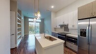 This contemporary single-floor residence showcases a thoughtfully designed open-concept layout with clean lines and modern finishes throughout. The kitchen serves as a centerpiece of the living space, featuring a large central island with a quartz countertop and undermount sink, accented by stylish pendant lighting. White upper cabinetry is paired with textured grey-toned lower cabinets, complemented by a classic subway tile backsplash with a decorative mosaic accent. A full suite of stainless steel appliances — including a French door refrigerator, electric range, and built-in microwave — provides both function and visual appeal. Rich hardwood-style flooring flows throughout the main living areas, leading to a sliding glass door that opens to an outdoor balcony, filling the space with natural light. A dedicated laundry area accommodates a full-size washer and dryer alongside built-in shelving for additional storage. The bedroom features plush carpeting and generous windows that invite ample daylight. The bathroom offers a dual-sink vanity with quartz countertops, warm-toned wood cabinetry, a large mirror, and direct access to a spacious walk-in closet. The overall ambiance is bright, clean, and move-in ready.