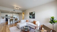 Located in Natura Gardens in Hialeah, Florida, this single-floor residence combines contemporary design with everyday functionality. The open-concept living area is bright and welcoming, featuring warm wood-tone flooring, neutral-toned walls, and a large window fitted with horizontal blinds that invite abundant natural light. The living space comfortably accommodates a sectional sofa, accent chairs, and a glass coffee table, creating an inviting gathering area.

The kitchen is sleek and modern, outfitted with white cabinetry, light quartz countertops, and a full suite of stainless steel appliances including a refrigerator, electric range, and over-the-range microwave. A breakfast bar with seating provides a casual dining option, and an in-unit laundry area is conveniently situated nearby.

The primary bedroom is serene and well-proportioned, offering access to a walk-in closet and featuring warm lighting and neutral finishes. A second bedroom provides additional flexibility, brightened by a window with outdoor views and accented by contemporary wall art. The bathroom is cleanly finished with marble-style tile in the tub-shower combination, a white vanity, and wood-look flooring. Throughout, the unit maintains a cohesive, modern aesthetic with thoughtful detailing.