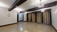 This single-floor space showcases a refined blend of modern and industrial design elements. The most commanding feature is a floor-to-ceiling textured accent wall finished in layered tones of bronze and brown, creating a dramatic focal point that anchors the main open area. Polished terrazzo flooring extends throughout, reflecting light and adding a sense of depth and elegance to the space.

Dark, structural ceiling beams run horizontally across the room, providing strong visual contrast against the lighter ceiling and walls. Suspended from the ceiling are multiple geometric chandeliers with a contemporary crystal-inspired design, casting warm, ambient light throughout and reinforcing the upscale character of the space.

The layout is open and versatile, with a corridor visible toward the rear that leads to additional areas, hinting at further utility beyond the main room. Dark door frames and base trim complement the bold beam work, maintaining a cohesive, sophisticated color palette. The overall ambiance is polished and professional, well-suited for a commercial lobby, event space, or public gathering area.