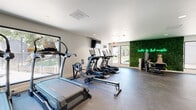 This fitness center is a thoughtfully designed, single-floor facility built to support a well-rounded workout experience. The space is outfitted with an extensive selection of cardio equipment, including TRUE-brand treadmills, elliptical trainers, StairMaster steppers, and stationary bikes, all arranged to maximize open floor space and ease of movement. A dedicated strength training area features a multi-functional cable machine, weight bench, and a fully stocked medicine ball and free weight rack, providing options for both functional and traditional resistance training. The flooring throughout is a durable dark rubber material suited for high-impact exercise. Recessed lighting keeps the interior bright and evenly lit, while large windows along multiple walls invite abundant natural light and offer views of an outdoor pool area and surrounding greenery. A wall-mounted flat-screen television is positioned near the entrance for entertainment during workouts. One of the most distinctive design elements is a full living green wall adorned with a glowing neon sign reading "hustle for that muscle," adding a motivating and modern aesthetic focal point to the space. A glass-paned door provides direct access to the outdoor grounds, further connecting the interior to the natural surroundings.