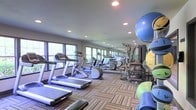 This fitness center is designed to support a comprehensive workout experience within a single, thoughtfully organized floor. The space is outfitted with a robust selection of cardio equipment, including multiple treadmills and elliptical machines, arranged in orderly rows to maximize accessibility and flow. A full multi-station strength training machine anchors the exercise area, complemented by a collection of stability balls and yoga mats for flexibility and functional training.

Large windows line the walls, inviting ample natural light and providing views of the surrounding greenery, creating an energizing atmosphere for workouts. Recessed ceiling lighting supplements the natural light, ensuring the space remains well-lit at all times of day. A floor-to-ceiling mirror runs along one wall, visually expanding the room and allowing users to monitor their form during exercise.

The flooring consists of durable, patterned carpet tiles in muted tones, offering both comfort underfoot and a clean, professional aesthetic. The overall ambiance is practical yet inviting, with a layout that balances open movement areas with organized equipment stations, making the fitness center suitable for a wide range of exercise routines and fitness levels.