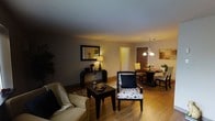 This well-appointed single-floor residence offers a seamless blend of comfort and style. The open-concept living and dining area serves as the heart of the home, unified by rich hardwood flooring that runs throughout and contributes to the space's warm, cohesive feel. The living area is thoughtfully arranged with a plush sofa, a patterned accent chair, and a dark wood coffee table, centered around a decorative area rug. A classic console table adorned with a table lamp and decorative accents anchors one wall, accompanied by a framed mirror that adds depth and reflects warm light across the room. Transitioning naturally into the dining area, a chandelier with elegant detailing hangs above a round dining table surrounded by matching chairs, creating an intimate setting for meals. Warm, ambient lighting throughout enhances the inviting atmosphere of the space. A doorway leads further into the home, suggesting additional private rooms beyond. The overall ambiance is cozy yet refined, making it well-suited for both relaxed everyday living and comfortable entertaining.