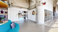 Located at 800 19th Ave S in Nashville, this expansive single-floor commercial space delivers a compelling blend of industrial architecture and contemporary design. Exposed concrete columns, concrete masonry block walls, and open ceilings with visible ductwork and conduit establish a raw industrial aesthetic, while thoughtfully curated furnishings and vibrant accent colors create a polished, functional environment throughout.

Floor-to-ceiling windows run along a significant portion of the perimeter, flooding the interior with abundant natural light and framing panoramic views of the Nashville skyline. Polished concrete floors extend through the primary common areas, complementing the structural materiality of the space.

The layout accommodates a variety of uses, including open collaborative lounges featuring bold, circular seating arrangements, a well-appointed kitchen and dining area with a vivid blue island and open shelving, and dedicated office zones with carpet tile flooring and track lighting. Private office suites with glass-front walls maintain visual connectivity while providing acoustic separation. Additional flex rooms and enclosed phone booths support focused work. Curated artwork adorns the walls, contributing to the space's creative, dynamic character.
