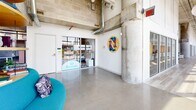 Located at 800 19th Ave S in Nashville, this expansive single-floor commercial space delivers a compelling blend of industrial architecture and contemporary design. Exposed concrete columns, concrete masonry block walls, and open ceilings with visible ductwork and conduit establish a raw industrial aesthetic, while thoughtfully curated furnishings and vibrant accent colors create a polished, functional environment throughout.

Floor-to-ceiling windows run along a significant portion of the perimeter, flooding the interior with abundant natural light and framing panoramic views of the Nashville skyline. Polished concrete floors extend through the primary common areas, complementing the structural materiality of the space.

The layout accommodates a variety of uses, including open collaborative lounges featuring bold, circular seating arrangements, a well-appointed kitchen and dining area with a vivid blue island and open shelving, and dedicated office zones with carpet tile flooring and track lighting. Private office suites with glass-front walls maintain visual connectivity while providing acoustic separation. Additional flex rooms and enclosed phone booths support focused work. Curated artwork adorns the walls, contributing to the space's creative, dynamic character.