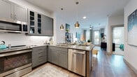 This single-floor residence offers a thoughtfully designed open-concept layout that seamlessly connects the kitchen, dining, and living areas. The kitchen is a standout feature, outfitted with sleek gray shaker-style cabinetry, granite countertops, a classic white subway tile backsplash, and a full suite of stainless steel appliances including a range, microwave, dishwasher, and refrigerator. A peninsula island with pendant lighting above provides additional prep space and casual seating for multiple bar stools. Luxury vinyl plank flooring runs throughout the main living areas, adding warmth and durability. The living room is bright and welcoming, with large windows dressed in patterned curtains that allow generous natural light and access to an outdoor area via a glass door. A ceiling fan adds comfort overhead. The space includes two bedrooms, each carpeted for comfort, with multiple windows offering natural light and outdoor views. The primary bathroom features dual granite-topped vanities with gray cabinetry, large mirrors, and a tub-shower combination. Additional storage is provided through closets with built-in shelving and hanging rods. The overall aesthetic is contemporary and cohesive, with a neutral palette accented by modern fixtures and finishes throughout.