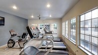 This fitness center is a dedicated single-floor workout facility thoughtfully designed to support an active lifestyle. The space is lined with large windows along multiple walls, flooding the interior with abundant natural light and providing views of the surrounding greenery, creating an open and motivating atmosphere. The light-toned walls, complemented by a subtle blue accent wall, contribute to a clean and energizing aesthetic. Recessed lighting and a ceiling-mounted fan ensure the space remains well-lit and comfortably ventilated during workouts. The facility is equipped with a row of treadmills and additional cardio machines, including stair steppers, all positioned to take advantage of the natural light from the windows. The flooring features a durable, textured surface suited for fitness use. A wall clock adds a practical touch for tracking workout sessions. The overall layout is efficient and spacious, allowing users to move freely between equipment. The combination of natural light, thoughtful design, and quality equipment makes this fitness center a functional and inviting space for daily exercise.