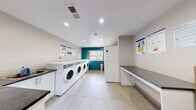 This shared laundry facility is thoughtfully designed to provide a clean, functional, and comfortable environment for everyday use. The room is finished with large-format light tile flooring and recessed ceiling lighting, creating a bright and welcoming atmosphere. A bold teal accent wall frames the central window, which allows natural light to fill the space and offers views of the surrounding neighborhood.

A row of modern front-loading washing machines lines one wall, each equipped with digital controls for ease of use. Stacked dryer units are positioned nearby, maximizing the room's capacity. Ample folding space is provided via wide countertops with dark stone-style surfaces, and a built-in utility sink adds practical convenience for hand-washing needs. White cabinetry beneath the counters offers additional storage.

A dedicated corner features a wooden bookshelf stocked with a variety of reading materials, alongside a cork bulletin board displaying notices and schedules — a thoughtful touch that encourages community engagement during wait times. Safety essentials, including a fire extinguisher and posted usage guidelines, are clearly visible near the entry door. The overall design balances utility with comfort, making this laundry room a well-appointed shared amenity.
