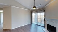 This single-floor space is designed with a clean, contemporary aesthetic that emphasizes openness and functionality. The main living area features smooth hardwood-style flooring in a cool gray-toned finish, lending a modern, cohesive look throughout. Vaulted ceilings add a sense of height and airiness to the room, enhancing the overall feeling of spaciousness.

A ceiling fan with a built-in light fixture is centrally mounted, providing both practical ventilation and ambient lighting. The walls are finished in a neutral light gray tone, offering a versatile backdrop suited to a variety of furnishing styles.

A standout feature of the space is the floor-to-ceiling sliding glass door, dressed with vertical blinds and leading to a private balcony. The balcony provides elevated outdoor views of surrounding greenery and the broader landscape beyond, allowing natural light to filter generously into the interior. White trim detailing along the walls and baseboards adds a crisp, finished appearance. Overall, the space conveys a bright, airy ambiance with thoughtful design details suited for comfortable everyday living.