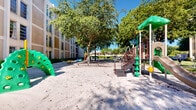 Located at 10255 Southwest 24th Street in Miami, FL, this outdoor playground offers a well-equipped recreational space set within a landscaped community environment. The play area features a variety of structures designed for children of different ages, including a multi-level climbing and sliding structure with a green canopy roof, a traditional metal slide with red staircase, a tunnel crawl arch, spring-mounted ride-on toys, and a freestanding swing set. The ground throughout the play area is covered in a sandy, loose-fill surface that provides cushioning beneath the equipment. Mature, broad-canopy trees are distributed throughout the space, casting generous shade across the sandy floor and creating a comfortable outdoor environment even under Miami's sunny skies. Wooden benches are positioned around the perimeter, offering seating for caregivers and visitors. A small community building with a covered roof is visible in the background, surrounded by well-maintained green lawns. The playground is flanked by multi-story residential buildings with large windows, contributing to a secure, community-centered atmosphere. Outdoor fitness equipment is also visible nearby, adding further recreational value to the space.