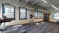 Located at 2054 Fulton Rd in Cleveland's Ohio City neighborhood, this fitness center occupies a single-floor space that blends industrial character with modern functionality. The facility is housed within a building that retains its original architectural charm, showcasing exposed structural beams on the ceiling, painted white brick walls, and rows of large multi-pane windows framed in dark trim. These windows allow generous amounts of natural light to fill the workout area while offering views of the surrounding neighborhood.

The fitness floor is spacious and open, with durable patterned carpet tile throughout. The cardio zone is well-appointed with multiple TRUE-brand treadmills, elliptical machines, and a stationary bike, all positioned to take advantage of the natural light streaming through the windows. A power tower provides options for bodyweight training, while a cable machine and multi-use weight bench support strength-focused workouts. A full dumbbell rack and a wall-mounted television are positioned alongside a large mirror, creating a dedicated free weights and functional training area. Stability balls in assorted sizes are stored on a wall-mounted rack nearby.

The space also features a glass-door enclosure separating a section of the gym, exposed structural columns with a raw industrial finish, and linear pendant lighting that supplements the natural illumination throughout. Overall, the facility offers a clean, well-organized environment suited for a comprehensive workout experience.