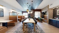 This thoughtfully designed single-floor space functions as a modern coworking lounge and café, blending professional functionality with inviting, café-inspired aesthetics. The layout is divided into several distinct seating zones, each offering a different atmosphere suited to various work styles and group sizes.

At the heart of the space sits a large central conference table surrounded by sleek black swivel chairs, anchored by a bold geometric-patterned area rug. A dramatic cluster of pendant lights in varying shapes and finishes hangs above, drawing attention to a richly textured wood-paneled accent wall and a distinctive painted ceiling that adds architectural depth.

Along one side, a row of semi-private booth seating features warm wood paneling and frosted glass dividers, providing quieter nooks ideal for focused work or smaller conversations. A separate intimate corner nook with plush teal velvet chairs, a round table, and a wall-mounted display screen offers a cozy spot for informal meetings, accented by artistic patterned wallpaper and a decorative globe light fixture.

The coffee bar area is a standout feature, showcasing bold signage, dark cabinetry, a full beverage station, and an adjacent built-in bench seating nook. Natural light filters throughout via large windows, while wood-look tile flooring and layered textures of brick, stone, and reclaimed wood create a warm, cohesive ambiance throughout the entire space.