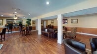 Located at 10580 Hammerly Blvd in Houston, Texas, this single-story community clubhouse offers a well-appointed gathering space designed for comfort, socializing, and events. The open floor plan is anchored by rich hardwood-style flooring that flows throughout, while warm two-tone walls in tan and terracotta create a welcoming ambiance.

The main lounge area is defined by elegant white structural columns and features an assortment of seating arrangements, including leather sofas, accent chairs, and high-top bar tables with upholstered stools — ideal for casual gatherings or relaxed conversation. Framed artwork adorns the walls throughout, adding character to the space.

A dedicated coffee and kitchenette area is outfitted with dark granite countertops, rich cabinetry, a stone tile backsplash, and a full-size refrigerator, providing convenient refreshment options for residents and guests.

The business center section includes a workstation with computer equipment, illuminated by recessed lighting and a ceiling fan. Large windows in this area invite natural light and offer views of the outdoor balcony area.

A standout feature is the raised performance stage at one end of the clubhouse, flanked by mounted speakers and a large-screen display — perfect for live entertainment, presentations, or community events. Comfortable lounge seating faces the stage, completing this versatile and well-equipped community space.