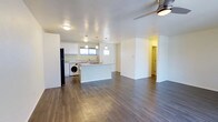This single-floor residence offers a thoughtfully designed open-concept layout that seamlessly connects the kitchen and living areas, creating a spacious and airy atmosphere. The kitchen is a standout feature, equipped with crisp white cabinetry, stone-style countertops, and a central island that provides additional workspace and serves as a natural gathering point. Pendant lighting hangs above the island, complementing the flush ceiling fixtures that illuminate the space. A black refrigerator anchors one end of the kitchen, while a washer unit is conveniently integrated into the kitchen zone, offering practical in-unit laundry access. Windows above the sink and near the kitchen area invite natural light, keeping the space bright and welcoming. Rich, dark hardwood-style flooring runs throughout the open area, adding warmth and contrast to the clean white walls and ceilings. A ceiling fan in the living area ensures comfort and air circulation. The layout flows into a spacious living zone with ample room for furnishing, and a hallway visible in the background suggests additional private rooms beyond. The overall ambiance is clean, modern, and functional.