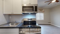 This compact single-floor unit offers a practical and efficiently designed layout comprising one bedroom and one bathroom. The main living area features light-toned laminate hardwood flooring and vertical shiplap-style wall paneling painted in a neutral gray tone, lending a clean, contemporary aesthetic throughout. A ceiling fan is installed overhead, contributing to climate comfort alongside wall-mounted air conditioning and baseboard heating units visible throughout the space.

The kitchen is equipped with stainless steel appliances, including a smooth-top electric range, an over-the-range microwave, and a refrigerator. White upper cabinetry provides storage, while dark-toned countertops and a stainless steel sink complete the functional cooking area. A window above the kitchen zone allows natural light to enter.

The bedroom is the largest area of the unit, featuring the same shiplap wall detailing, laminate flooring, a window with horizontal blinds, and baseboard heating. The layout is straightforward and open, maximizing usability within the available space.

The bathroom is finished with white subway tile walls, tile flooring, a white vanity with a framed mirror, a bathtub, and a baseboard heater. The overall design is clean and functional, prioritizing practicality throughout.