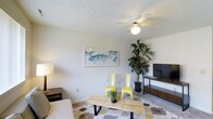 This two-story townhome offers a well-proportioned layout designed for comfortable everyday living. The main living area, visible in the image, features neutral-toned walls and a textured ceiling with a ceiling fan and integrated light fixture, providing both air circulation and ambient lighting. Carpet flooring with a patterned area rug adds warmth and texture to the space. The living room is thoughtfully arranged, with room for seating, an entertainment setup, and decorative accents including wall art and indoor greenery that contribute to a relaxed, coastal-inspired atmosphere. Natural light filters into the space, keeping the interior feeling open and bright. The two-story configuration of the townhome allows for a clear separation between the living areas on the main floor and private spaces on the upper level, a hallmark of townhome-style living. The overall ambiance is casual yet inviting, with a neutral color palette that provides a versatile backdrop for personalization. The space strikes a balance between functionality and comfort, making it well-suited for a variety of living arrangements.