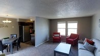 This single-floor space features an open-concept layout that connects the living area to the kitchen, creating a cohesive and functional living environment. The living area is finished with wall-to-wall carpeting in a neutral gray tone, providing a comfortable and practical surface underfoot. Two windows along the far wall allow natural light to fill the room, brightening the space and offering outside views. The textured popcorn ceiling adds a classic finish overhead throughout the unit.

The kitchen, visible from the living area, is equipped with dark wood cabinetry, a stainless steel refrigerator, and a stove, offering a practical cooking space. A countertop bar area provides a natural transition between the kitchen and living space, making it ideal for casual dining or meal preparation. The gray walls throughout maintain a neutral, clean aesthetic that complements both the carpeted flooring and the darker kitchen finishes. Overall, the space presents a straightforward, functional layout well-suited for everyday living.