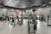 This fitness center offers a generously sized, single-floor workout facility designed to support a full range of exercise activities. The space is defined by a striking architectural ceiling treatment — a dark navy tray ceiling with linear LED lighting and ceiling fans — set against crisp white walls and light wood-look tile flooring, creating a modern, clean aesthetic throughout.

The main workout area is open and thoughtfully arranged to maximize usable floor space. It is outfitted with a comprehensive selection of Cybex strength-training machines, including cable machines, seated press stations, and isolateral equipment, positioned to allow comfortable circulation between stations. A dedicated free weight area features a multi-tier dumbbell rack paired with an adjustable weight bench, along with a coat hook nook nearby. Cardio equipment, including treadmills, ellipticals, and stair climbers, lines the perimeter.

Large windows and glass doors on multiple walls flood the interior with natural light and provide views of the surrounding outdoor area, enhancing the bright and energetic atmosphere. Wall-mounted televisions add convenience for users during workouts. The facility benefits from a secondary room that offers additional equipment space and direct outdoor access through glass double doors, further connecting the interior to its exterior surroundings.