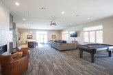 This well-appointed single-floor community space is thoughtfully designed to accommodate a variety of social and functional needs. The kitchen area features warm wood-grain cabinetry, a teal island with a crisp white countertop, and seating for several guests on bold navy blue bar stools. Stainless steel appliances and a tiled backsplash complete the polished, modern look, while pendant lighting adds an inviting ambiance overhead.

Adjacent to the kitchen, a spacious lounge area offers generous seating with neutral-toned sectional sofas, a wall-mounted flat-screen television, and large windows that fill the room with natural light. A cozy corner features a linear fireplace set against tile surround, complemented by warm yellow accent walls and a classic leather armchair, creating an intimate retreat within the larger space.

The game area includes a full-size billiards table and additional lounge seating, making it ideal for recreation and socializing. Toward the entrance, a meeting and dining zone provides multiple tables and chairs alongside glass double doors that open directly to the exterior, offering convenient access and views of the surrounding landscape. Consistent carpeted flooring and recessed lighting unify the space throughout.