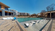 Located at 305 Prine Pl in Charlotte, NC, this outdoor amenity space is designed to deliver a resort-caliber experience. At its centerpiece is a generously sized swimming pool with crystal-clear blue water, outfitted with in-water lounge chairs that invite residents to relax partially submerged in the shallow entry area. A row of additional chaise lounges lines the pool deck, providing ample space for sunbathing under the open Carolina sky. An ADA-accessible pool lift ensures inclusive access for all users.

Adjacent to the pool, a covered outdoor lounge area features a warm wood-paneled ceiling with recessed lighting, supported by bold blue structural columns. This semi-open pavilion is furnished with plush, cushioned sofas and accent chairs styled with navy and orange throw pillows, alongside concrete-topped side tables — creating a comfortable, design-forward gathering space with direct views of the pool.

The outdoor dining area extends from this pavilion, equipped with modern slatted dining tables and chairs, as well as a wall-mounted outdoor television — ideal for entertaining. Brick-patterned pavers throughout the space add a refined, cohesive aesthetic. Glass doors bearing the community's "Live + Thrive" motto connect the outdoor space to the interior clubhouse, seamlessly blending indoor comfort with outdoor living.