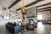 This thoughtfully designed single-story space blends rustic and contemporary elements throughout its open, flowing layout. Exposed dark wood beams cross the ceiling above wide-plank hardwood flooring, establishing a warm architectural rhythm that carries throughout the main lounge area. A dramatic floor-to-ceiling patterned tile surround frames the central fireplace, serving as the room's defining focal point. Woven pendant light fixtures hang overhead, complementing the eclectic mix of leather sofas, curved wood rocking chairs, and layered seating arrangements that define distinct conversation zones.

A gallery wall near the entry features a curated collection of framed artwork alongside an oval black mirror, adding a creative, editorial quality to the space. Multiple large windows fitted with roller shades admit generous natural light while offering views of the surrounding outdoor environment.

The kitchen area is distinctly styled with sage green cabinetry topped with dark countertops, open industrial-style shelving mounted against botanical-patterned wallpaper, and an intricate geometric tile floor. A beverage cooler and ample counter space make it well-suited for entertaining. A separate dining area with a rustic wood table and casual seating rounds out the space, reinforcing its communal and welcoming character.