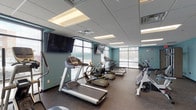 Located at 7856 S State Rt 48 in Maineville, Ohio, this single-floor fitness facility is designed to provide a comprehensive workout environment. The space is defined by its teal-painted walls, drop-tile ceilings with recessed lighting, and durable rubber-composite flooring throughout — a practical and clean aesthetic well-suited for an active facility.

The cardio zone is well-equipped, featuring multiple treadmills, elliptical trainers, a rowing machine, and stationary spin bikes arranged to take full advantage of the large floor-to-ceiling windows lining the perimeter. These expansive windows flood the space with natural light and offer views of the surrounding exterior, creating an open and energizing atmosphere. Multiple wall-mounted flat-screen televisions are positioned throughout for entertainment during workouts.

A dedicated strength and functional training area houses a Precor cable machine system, a recumbent bike, stability balls, and a variety of resistance bands and suspension straps mounted along the wall. This section provides ample open floor space for stretching, functional movements, and rehabilitation-style exercises.

The facility is thoughtfully laid out to separate cardio and strength zones while maintaining an open, accessible feel. With its abundant natural light, modern equipment, and clean design, this fitness space delivers a well-rounded and functional environment for users of all fitness levels.