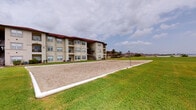 Located at 1802 Ennis Joslin Rd in Corpus Christi, Texas, this fitness center is a well-appointed, single-floor facility designed to support a wide range of training styles. The interior is bright and modern, featuring crisp white walls, recessed lighting, and large windows that allow natural light to pour in while offering views of the lush surrounding landscaping, including tropical palm trees.

The gym is divided into functional training zones. A dedicated free weights area houses a full rack of dumbbells alongside a weight bench and squat/power rack with an extensive plate selection. A cable and multi-station machine provides versatility for resistance training, all reflected by floor-to-ceiling mirrors that enhance the sense of space. A turf-covered training zone features a heavy punching bag, suspension trainers, a pull-up rig, medicine balls, and plyometric boxes — ideal for functional and high-intensity workouts. Motivational signage with phrases like "GRIT," "HUSTLE," and "EXECUTION" reinforces the facility's performance-focused atmosphere. Rubber flooring throughout ensures safety and durability.

Outside, a full-size sand volleyball court sits adjacent to the building, surrounded by well-maintained green lawns — a standout amenity perfect for recreational activity and community engagement.
