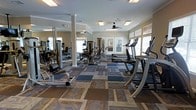 This fitness center offers a comprehensive workout environment designed to accommodate a variety of exercise routines. The space is outfitted with a broad selection of equipment, including multiple treadmills, cable machines, and strength-training stations, all thoughtfully arranged to allow for comfortable movement throughout the floor. Large windows along the perimeter flood the room with natural light, creating an open and energizing atmosphere. Ceiling fans are mounted overhead to promote air circulation and comfort during workouts. Full-length mirrors line one wall, enhancing the sense of spaciousness while allowing users to monitor their form. The flooring consists of a patchwork of durable, multi-toned carpet tiles that define different zones within the space. Structural columns are integrated into the layout without disrupting the overall flow of the room. Crown molding along the ceiling adds a subtle architectural detail, lending a polished finish to the space. The combination of ample natural light, well-maintained equipment, and thoughtful design makes this fitness center a functional and inviting facility for daily use.