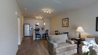 This single-floor residence offers a thoughtfully designed open-concept layout that seamlessly connects the main living areas. The kitchen is outfitted with white cabinetry, granite-style countertops, stainless steel appliances including a full-size refrigerator and microwave, and a breakfast bar with seating for two. Pendant lighting above the bar and a coordinating track light fixture add a modern touch to the space. Adjacent to the kitchen, a compact dining area provides room for a table and chairs, making it well-suited for everyday meals or casual entertaining. The living room flows naturally from the dining space, featuring neutral-toned upholstered furniture and warm hardwood-style flooring that runs throughout the unit, lending a cohesive and inviting atmosphere. Recessed and track lighting ensures the space remains bright and well-illuminated. The neutral wall tones and clean architectural lines give the interior a contemporary yet welcoming character, making it a practical and comfortable environment suited for a variety of lifestyles.