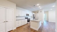 This thoughtfully designed 3-bedroom, 2-bathroom single-story home offers a clean, modern aesthetic with a functional layout. The open-concept kitchen and living area serves as the heart of the home, showcasing crisp white shaker cabinetry that extends to the ceiling, granite countertops, and a full suite of stainless steel appliances including a range, microwave, and French door refrigerator. A central kitchen island with a built-in sink and granite surface provides additional prep space and casual seating. Warm-toned hardwood-style flooring flows throughout the main living areas, lending a cohesive and inviting feel. The three bedrooms are carpeted for comfort, each featuring ceiling fans and windows that welcome abundant natural light. The primary bedroom includes access to a walk-in closet equipped with multi-level shelving and hanging rods for organized storage. The two bathrooms continue the home's refined palette, featuring white cabinetry, granite countertops, and hardwood-style flooring. A hallway connects the rooms efficiently, with three closets distributed throughout for added convenience. The home's neutral tones, recessed lighting, and consistent finishes create a bright, clean ambiance throughout.