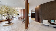 This single-floor amenity space is defined by its standout curved architectural centerpiece — a dramatic cylindrical wall clad in dark herringbone-patterned wood, flanked by vertical timber slat screens that segment the space into distinct yet interconnected zones. Large-format cream tile flooring flows throughout the open common areas, grounding the design in a clean, modern aesthetic.

The main lounge area is generously sized and bathed in natural light from expansive windows that frame views of palm trees and a resort-style outdoor area. Multiple seating arrangements — featuring plush sofas, ottomans, and accent chairs — are anchored by a vibrant, multicolored area rug and illuminated by striking geometric pendant chandeliers. A wall-mounted television above a sleek fireplace serves as a focal point.

A dedicated entertainment nook, wrapped in dark herringbone wall paneling, houses a curved leather sofa and a copper-framed television, offering a more intimate setting. Adjacent to this, a full billiards room with herringbone hardwood flooring centers around a full-size pool table beneath focused pendant lighting.

The well-appointed kitchen features rich wood cabinetry, white quartz countertops, an island with bar seating, and large windows overlooking the outdoor amenities — ideal for entertaining. Curated sculptural décor and ornamental trees add warmth and personality throughout.