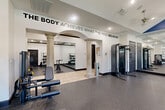 Located at 12430 Oxford Park Dr in Houston, TX, this fitness center offers a spacious, single-floor layout designed to support a full range of workout activities. The facility is divided into distinct functional zones, seamlessly connected by an elegant archway framed by decorative classical columns that lend a refined architectural character to the space.

The strength training area features a multi-station cable machine, a weight bench, and a dedicated free weights section with a full dumbbell rack positioned in front of floor-to-ceiling mirrors — allowing users to monitor their form during exercise. Adjacent to this zone, a cardio section houses multiple stationary cycling bikes and a stair climber, offering a well-rounded selection of equipment. Recessed lighting and ceiling fans are installed throughout, ensuring the space remains well-lit and ventilated.

The flooring transitions between durable rubber athletic matting in the workout zones and light tile in the hallway and entryway areas. A vaulted ceiling with a bold blue accent panel adds visual height and energy to the main training floor. The walls feature large-scale motivational typography, including the phrase "The Body Achieves What the Mind Believes," reinforcing the space's purpose-driven atmosphere. A glass-panel entry door allows natural light to filter into the hallway, creating a welcoming transition into the facility.