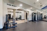 Located at 12430 Oxford Park Dr in Houston, TX, this fitness center offers a spacious, single-floor layout designed to support a full range of workout activities. The facility is divided into distinct functional zones, seamlessly connected by an elegant archway framed by decorative classical columns that lend a refined architectural character to the space.

The strength training area features a multi-station cable machine, a weight bench, and a dedicated free weights section with a full dumbbell rack positioned in front of floor-to-ceiling mirrors — allowing users to monitor their form during exercise. Adjacent to this zone, a cardio section houses multiple stationary cycling bikes and a stair climber, offering a well-rounded selection of equipment. Recessed lighting and ceiling fans are installed throughout, ensuring the space remains well-lit and ventilated.

The flooring transitions between durable rubber athletic matting in the workout zones and light tile in the hallway and entryway areas. A vaulted ceiling with a bold blue accent panel adds visual height and energy to the main training floor. The walls feature large-scale motivational typography, including the phrase "The Body Achieves What the Mind Believes," reinforcing the space's purpose-driven atmosphere. A glass-panel entry door allows natural light to filter into the hallway, creating a welcoming transition into the facility.