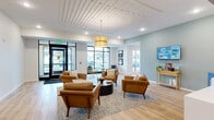 Located at 9900 56th Ave N in Plymouth, Minnesota, this single-floor community lobby and leasing center offers a thoughtfully designed welcome experience for residents and visitors alike. The main living area serves as an open, airy lounge anchored by a seating arrangement of warm cognac leather armchairs and a round coffee table atop a textured area rug. Light wood-plank flooring flows throughout, complementing the soft sage-green accent walls and crisp white surfaces that define the space's contemporary aesthetic. A statement drum pendant in brushed gold hangs from a coffered white ceiling, adding a refined focal point above the lounge. A sleek white reception desk occupies one side of the main area, complete with workstations and wall-mounted displays providing community information. Floor-to-ceiling black-framed windows and glass entry doors flood the lobby with natural light and offer views of the surrounding neighborhood. Curated abstract artwork lines the walls in the transitional hallway zones, lending an art-gallery quality to the circulation corridors. Directional signage in the hallways guides visitors toward the Fitness & Yoga Studio and the Clubroom & Sports Simulator, highlighting the range of amenities accessible from this central hub. The overall ambiance is polished yet approachable, blending modern design sensibilities with comfortable, functional furnishings.